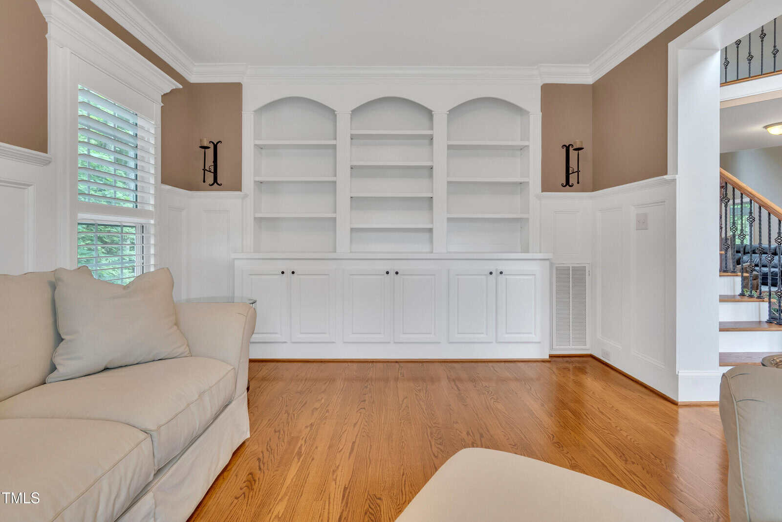 Bright living room with white built-in shelves and cabinets, light brown walls, hardwood floor, and beige couches in the foreground. A staircase is visible on the right side.