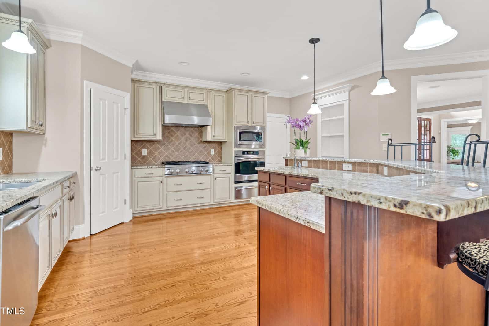 A spacious kitchen with wooden floors, beige cabinets, stainless steel appliances, granite countertops, pendant lights, and a central island with a raised bar area. Purple flowers decorate the countertop.