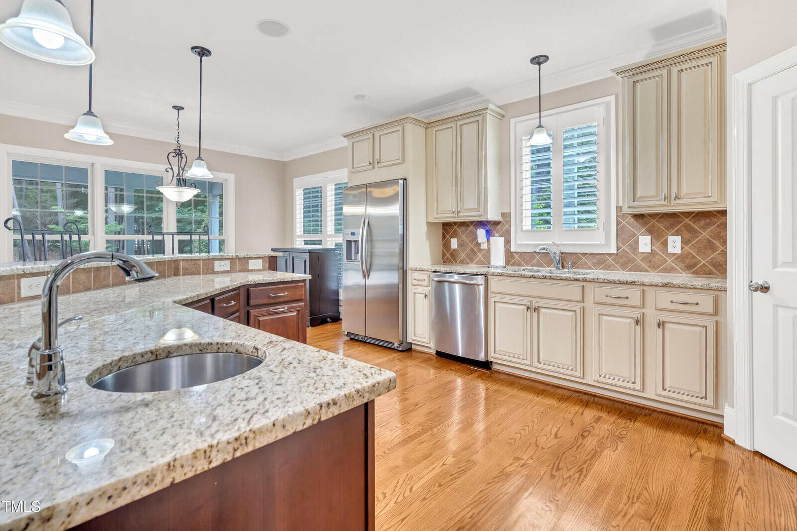 Spacious kitchen with granite countertops, stainless steel appliances, white cabinets, a center island with a sink, pendant lighting, tiled backsplash, and hardwood floors. Large windows provide natural light.