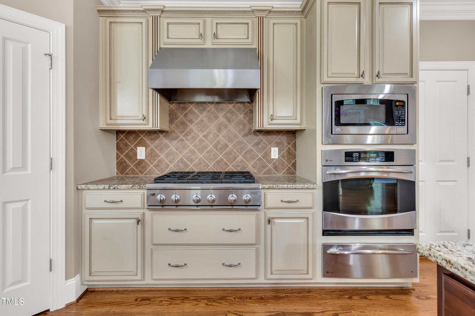 A kitchen with a gas cooktop and stainless steel range hood, tan cabinets, granite countertops, a built-in microwave, and an oven, with a brown tile backsplash and hardwood floor.