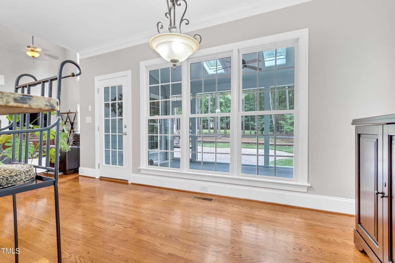 A dining area with hardwood floors, a hanging light fixture, large windows, a glass door, and views of a screened porch and trees outside. A dark cabinet stands against the right wall.