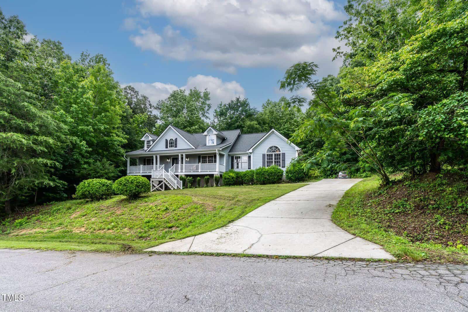 A large, gray house with a wraparound porch sits on a small hill, surrounded by lush green trees. A long, curved driveway leads up to the house from a quiet road under a partly cloudy sky.