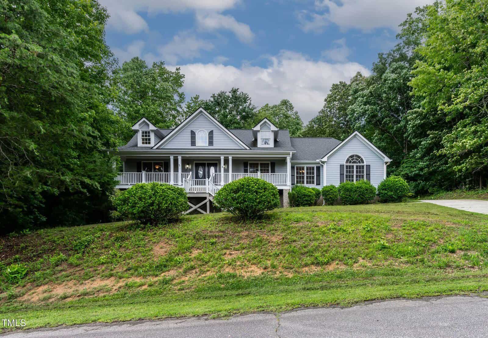 A light blue house with a large front porch, white railings, and multiple gables, surrounded by lush green trees and shrubs, on a grassy lawn with a driveway on the right.