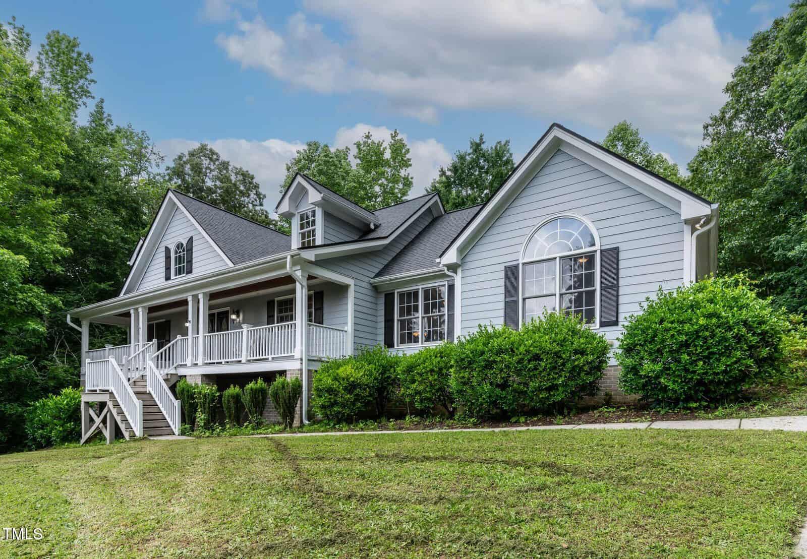 A light gray, two-story house with white trim, a large front porch, multiple windows, and a well-maintained lawn, surrounded by green trees under a partly cloudy sky.