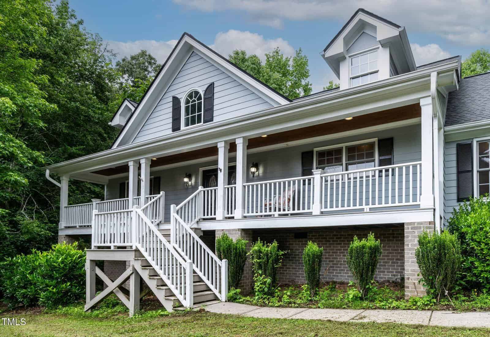 A gray, two-story house with white trim and a wide covered front porch featuring white railings and columns, surrounded by green shrubs and trees under a partly cloudy sky.