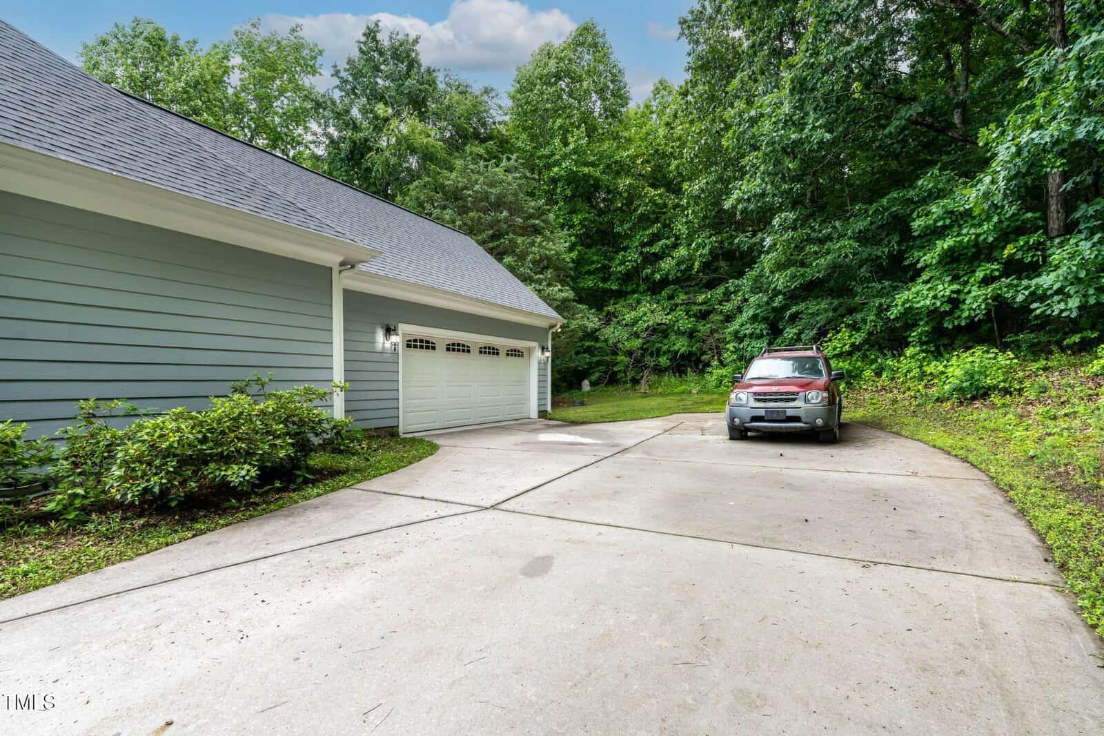 A silver SUV is parked on a wide concrete driveway next to a gray house with a three-car garage. Lush green trees and bushes surround the driveway and house.