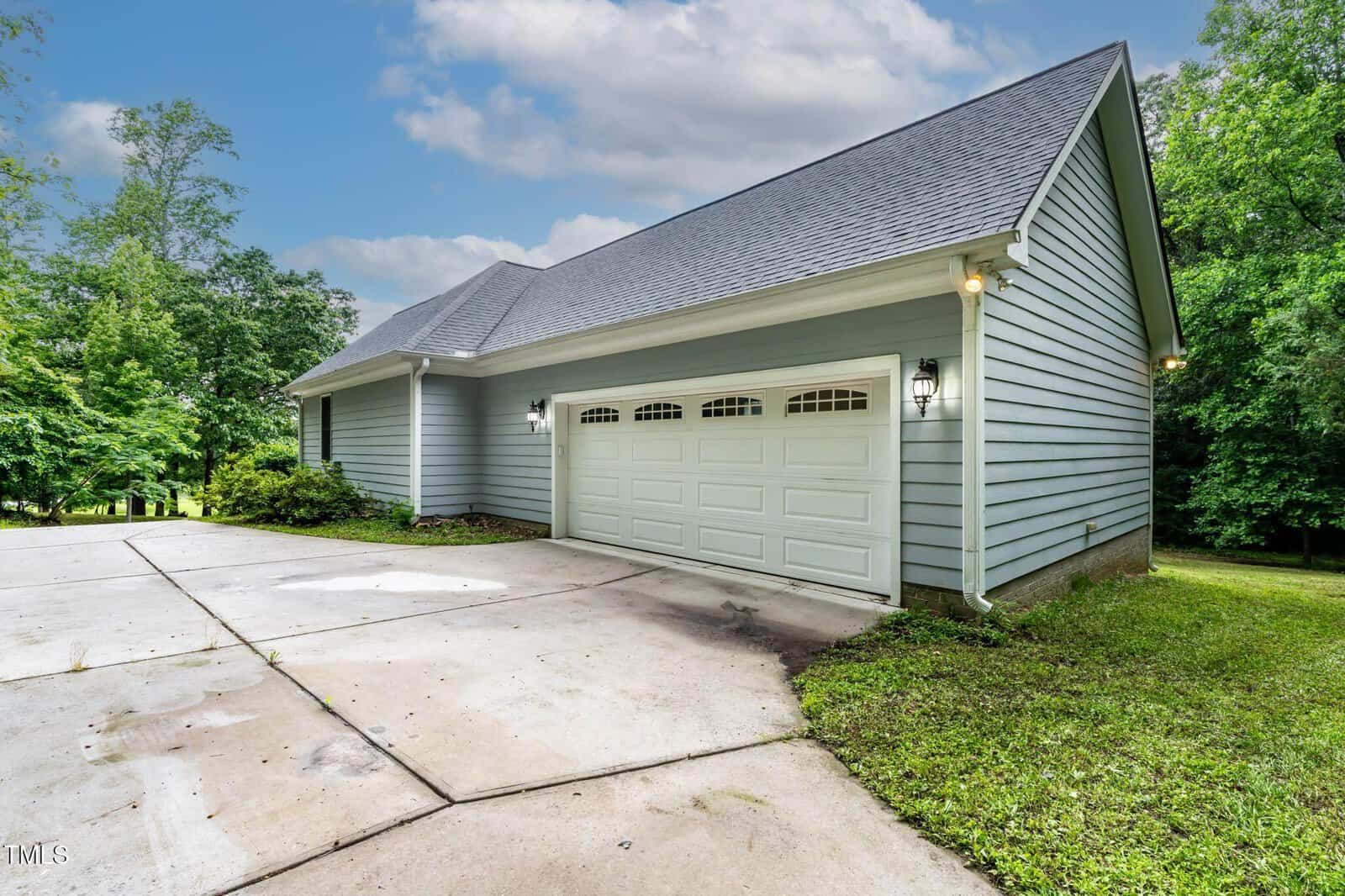 A light blue house with a two-car garage, white trim, and a large concrete driveway, surrounded by green trees under a partly cloudy sky.