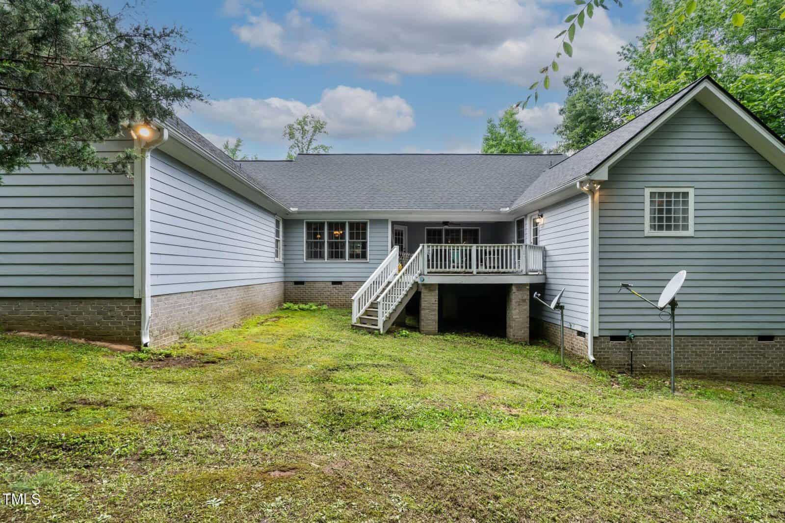 A light gray house with a dark roof, raised back porch, white railings, and stairs leading to a grassy backyard. Satellite dishes and basement windows are visible on the right side. The yard is bordered by trees.