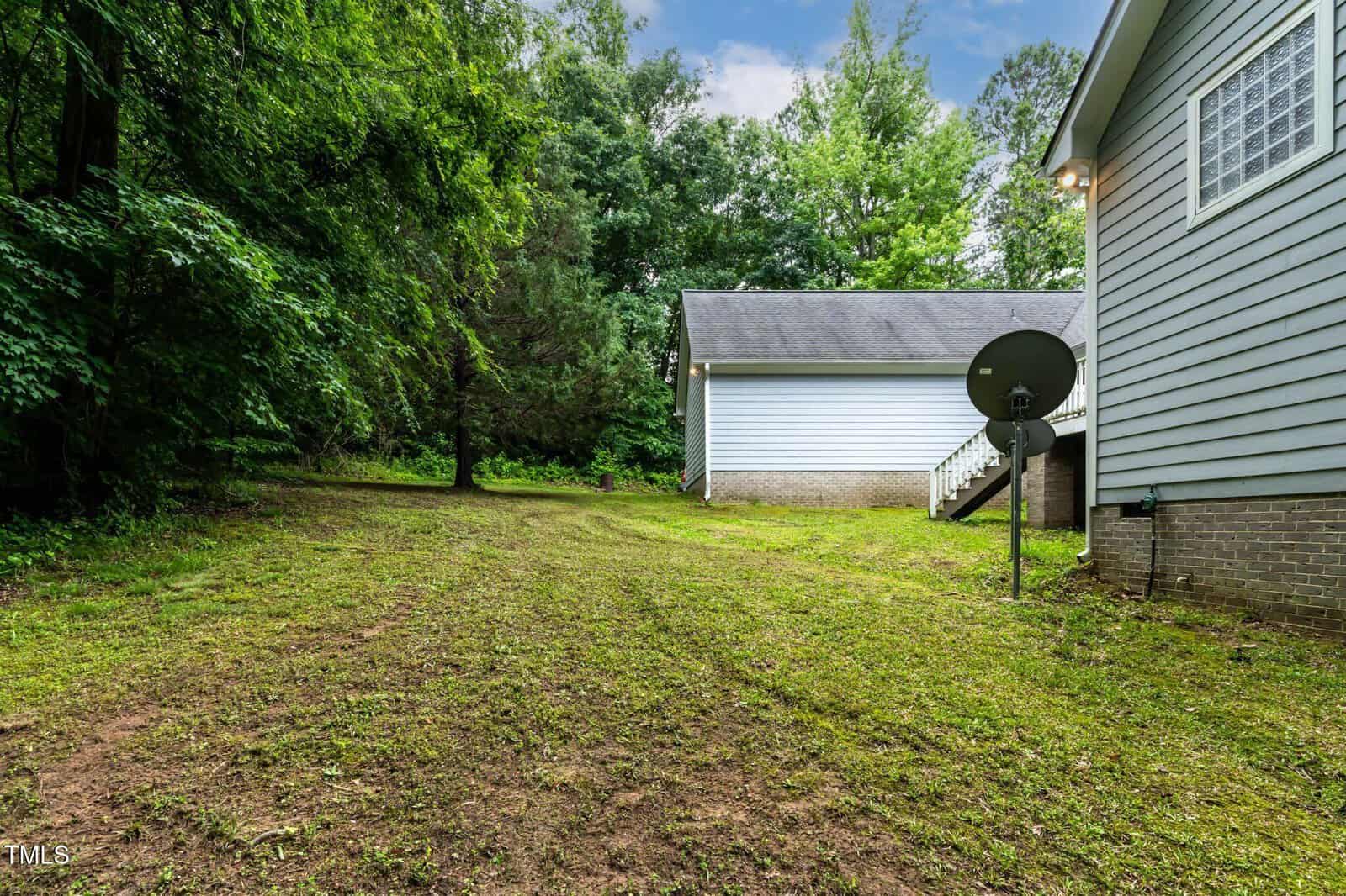 A grassy backyard with tall trees on the left, a light gray house with a satellite dish and stairs on the right, and another building in the background under a partly cloudy sky.