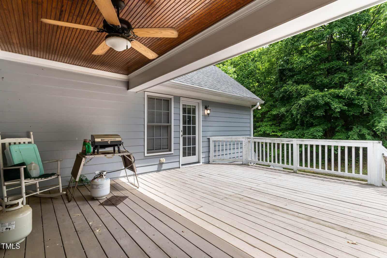 A covered wooden deck with a ceiling fan, grill, propane tanks, chairs, and a railing, attached to a gray house with glass doors and surrounded by green trees.