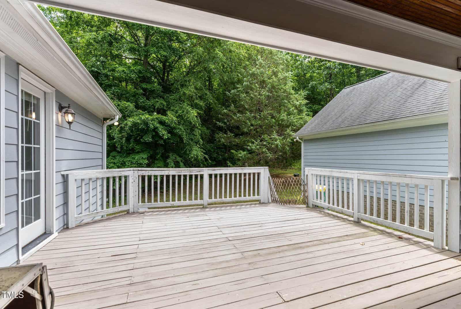 Spacious wooden deck with white railing, attached to a gray house with French doors, overlooking a green, tree-filled backyard and a detached gray garage.