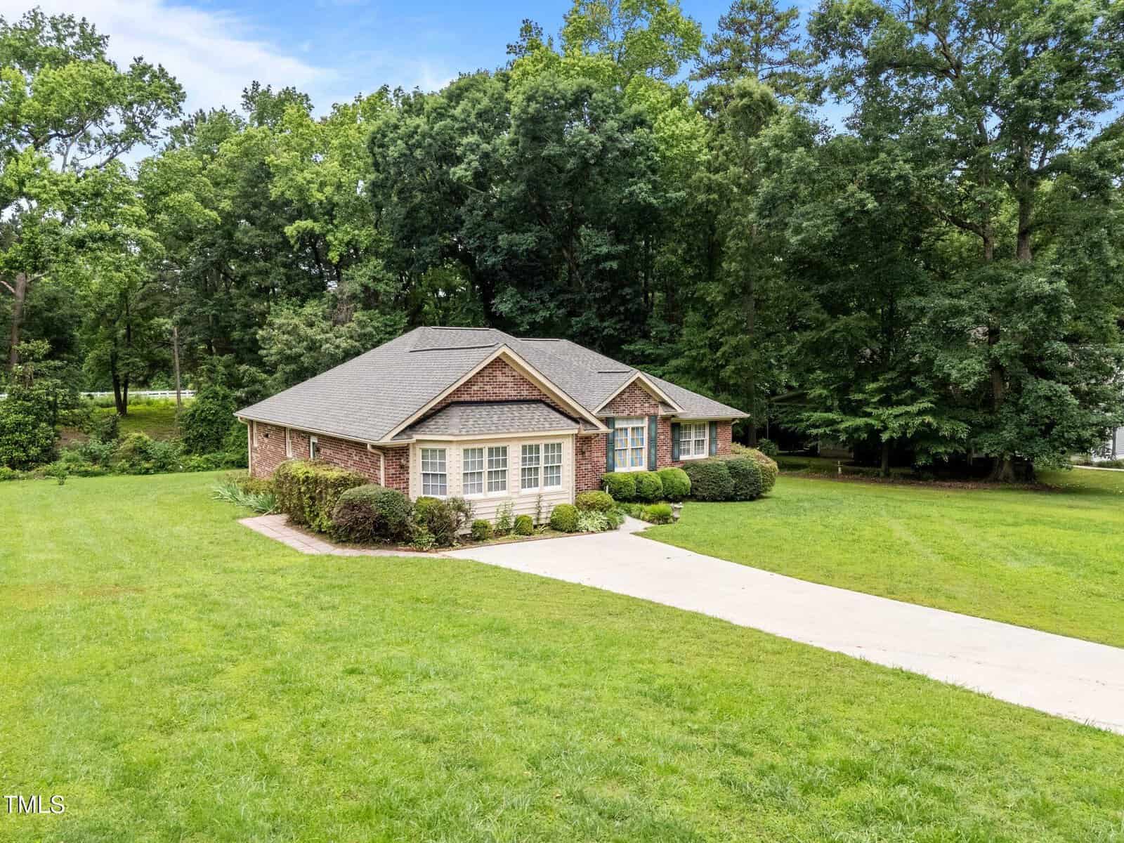 Single-story brick house with gray roof, white trim, and front shrubs, set on a large lawn with a concrete driveway and surrounded by mature trees. Blue sky is visible above the treeline.