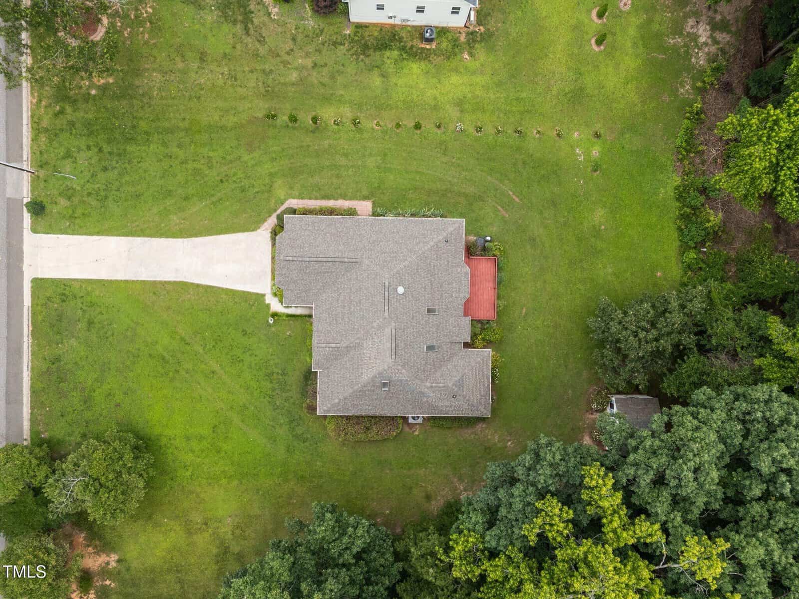 Aerial view of a single-story house with a gray roof, driveway, and red deck in a spacious, green yard. Trees border part of the property, and another structure is partially visible nearby.
