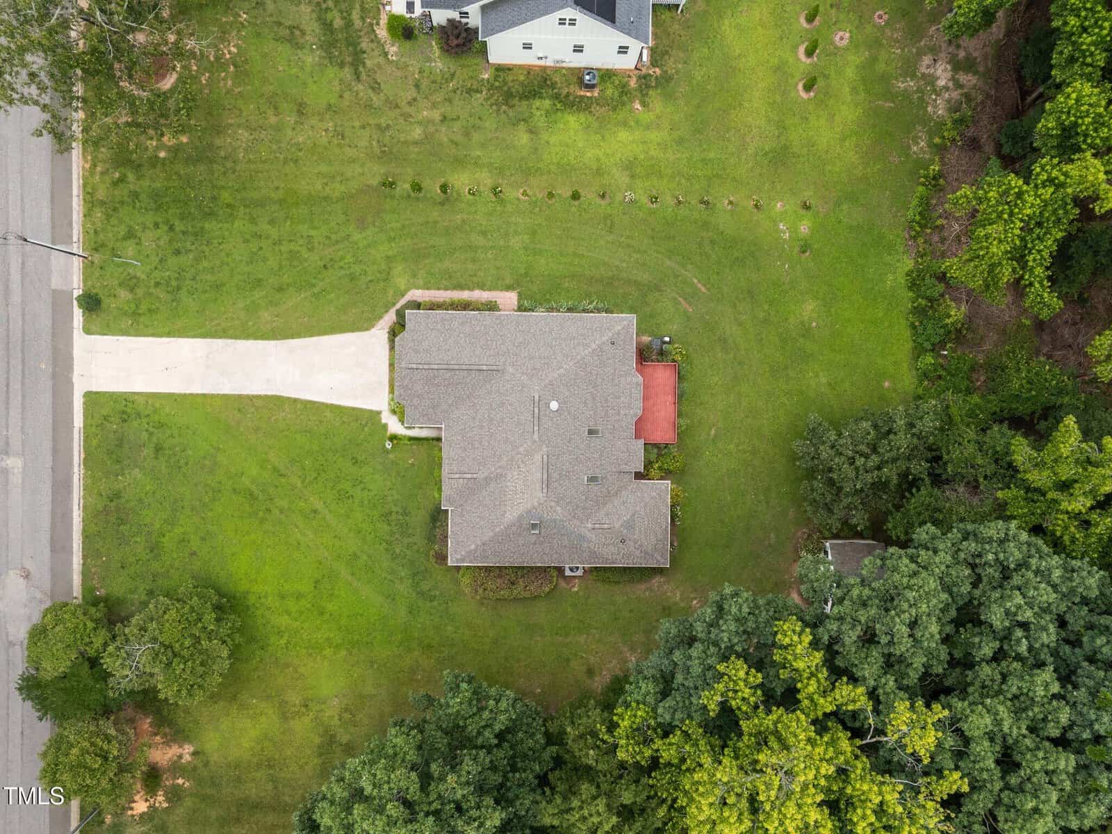 Aerial view of a single-story house with a gray roof, large green lawn, driveway leading to the street, and a red deck in the backyard. Trees and neighboring properties surround the house.