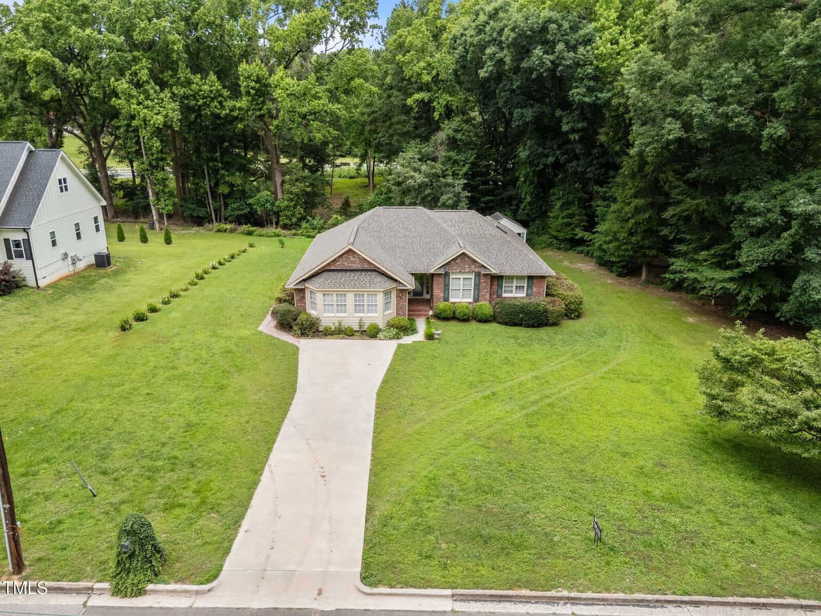 A single-story brick house with a gabled roof sits on a large, well-maintained lawn with bushes and trees. A concrete driveway leads from the street to the front entrance. The house is surrounded by greenery.