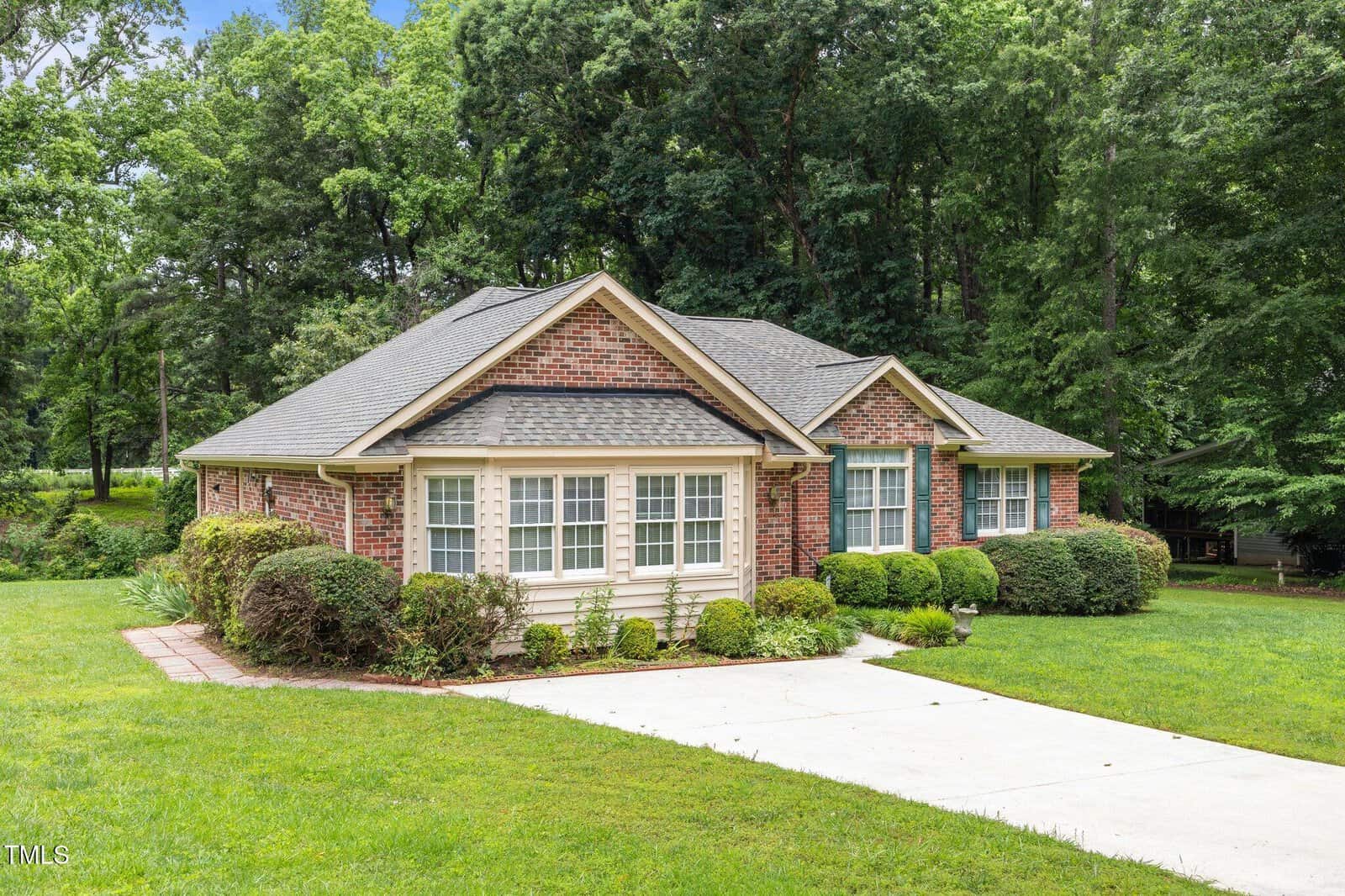 Single-story brick house with white-trimmed windows, surrounded by green shrubs and a lawn, set against a backdrop of tall trees. A curved driveway leads to the front.