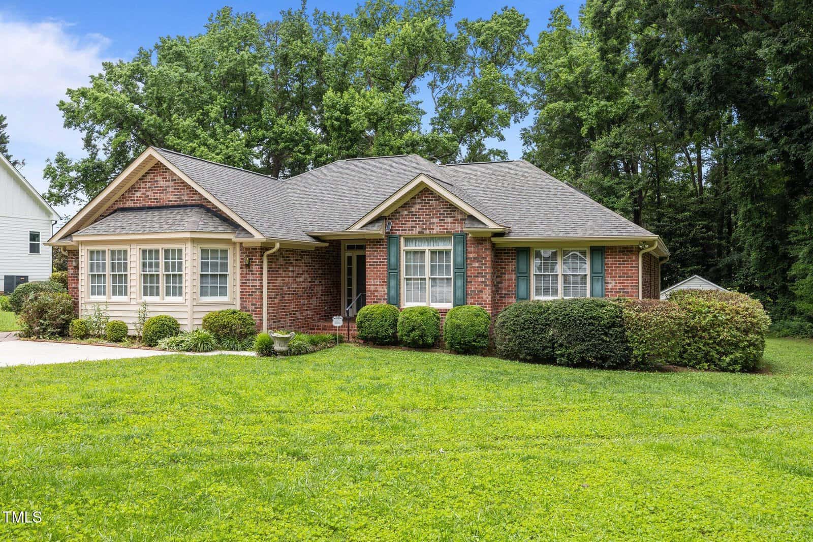 Single-story brick house with beige siding, gray roof, white trim, green shutters, shrubs along the front, and a well-kept lawn, surrounded by trees under a blue sky with clouds.