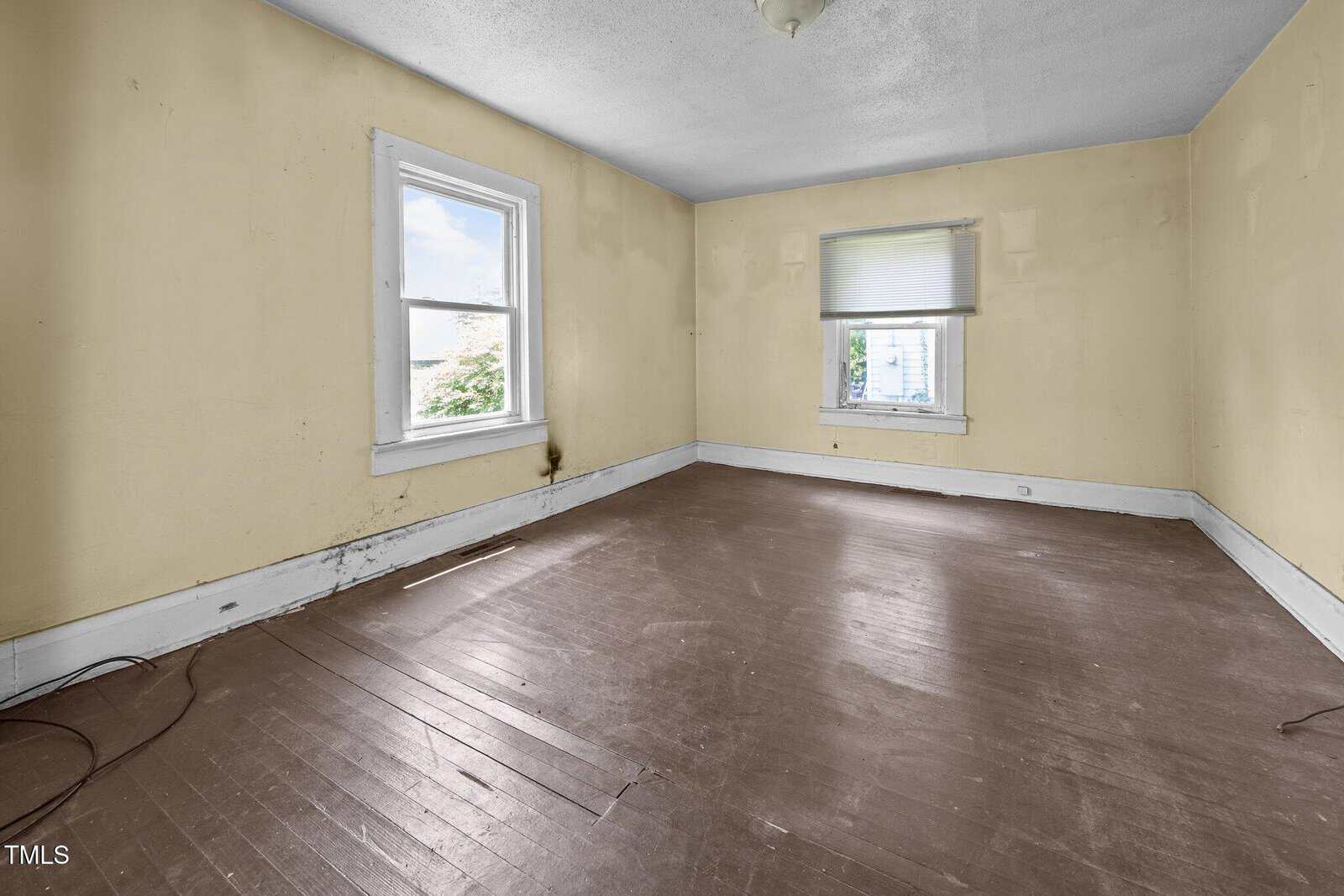 Empty room with yellowed walls, two windows with white trim, brown wooden floor, and white baseboards. One window has a partially closed blind; the room is well-lit with natural light.