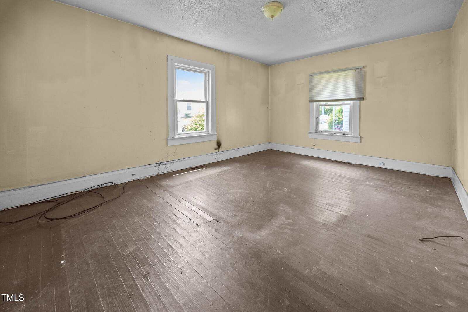 Empty room with beige walls, two windows with white trim, brown wooden floor, some scuff marks and stains on the walls and floor, a ceiling light fixture, and visible electrical cords along the baseboard.