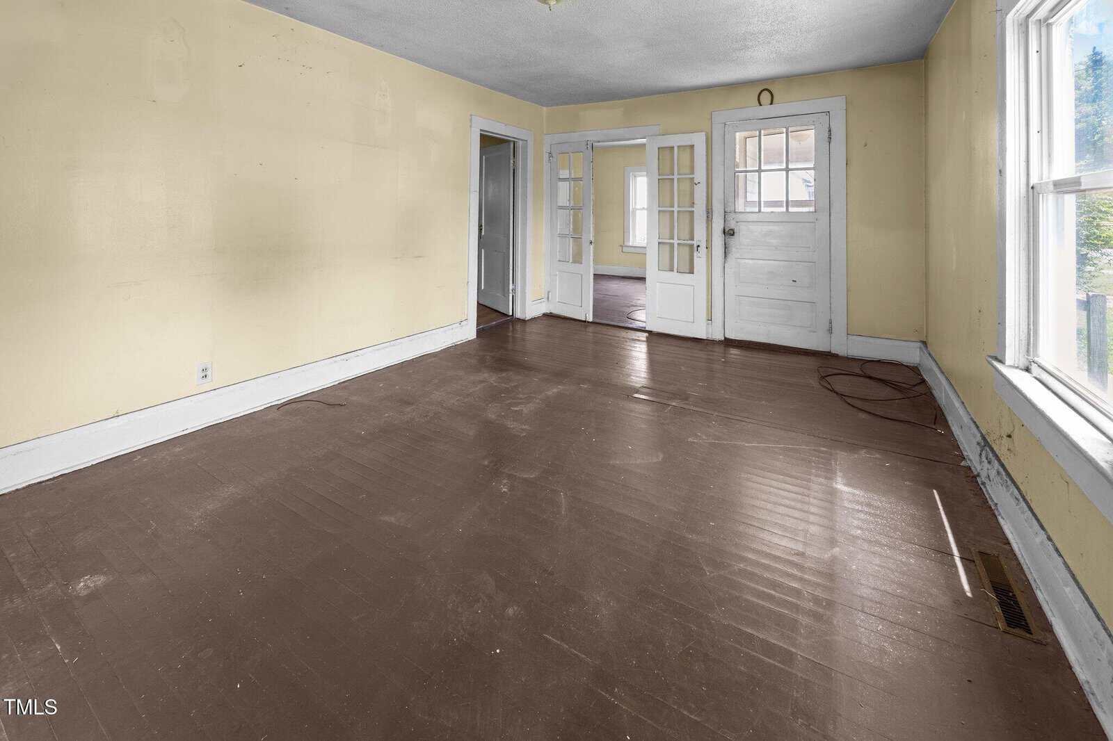 An empty room with brown painted wooden floors, pale yellow walls, two large windows on the right, and glass-paneled doors leading to another room. There is visible wear on the floor and some marks on the walls.