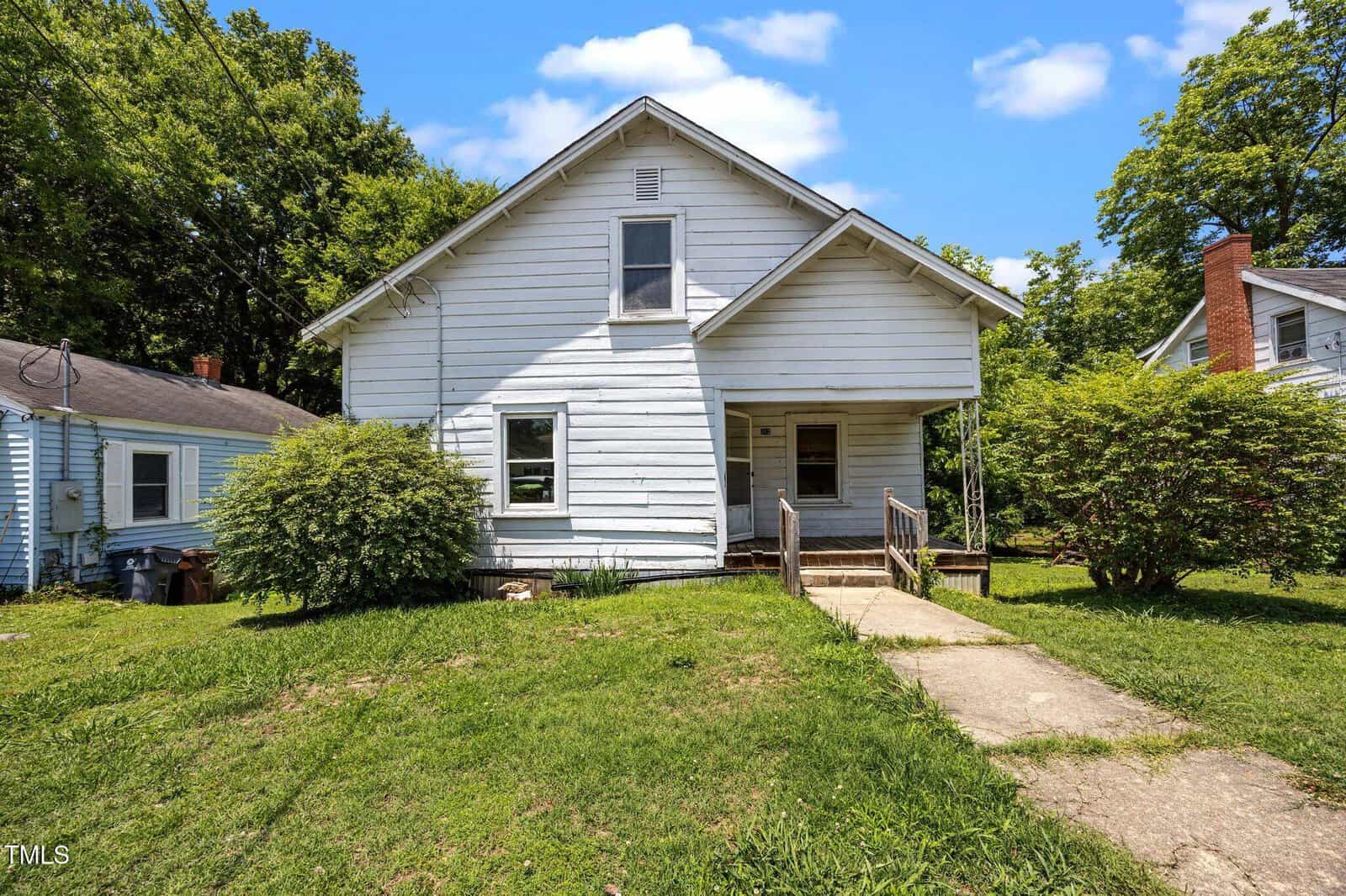 A white, two-story house with a sloped roof and small front porch, surrounded by grass, bushes, and trees. A concrete path leads from the sidewalk to the front door. The sky is blue with a few clouds.