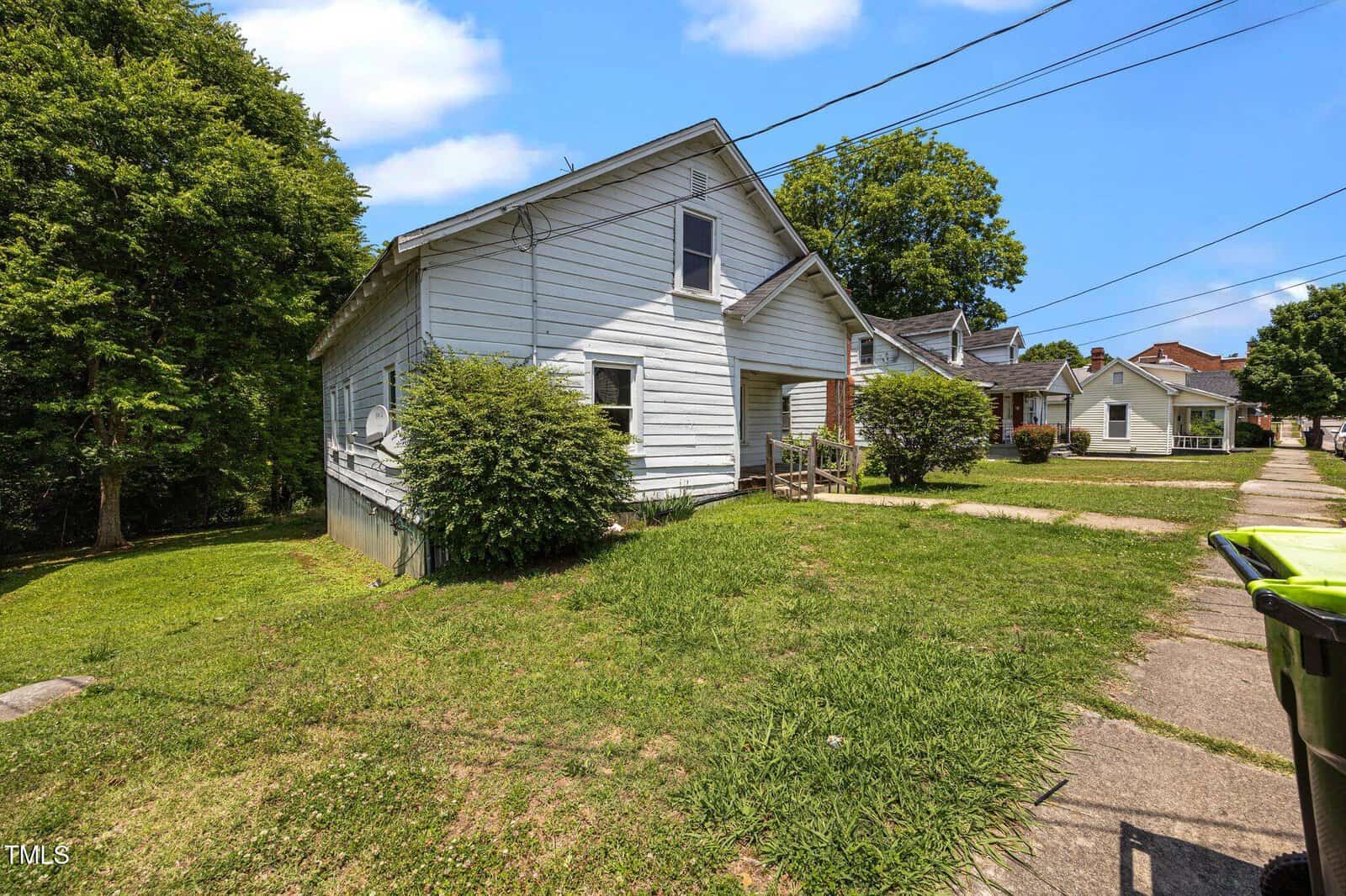 A white, single-story house with a sloped roof sits on a grassy yard, bordered by a sidewalk and driveway. Trees and neighboring houses are visible in the background under a sunny, partly cloudy sky.