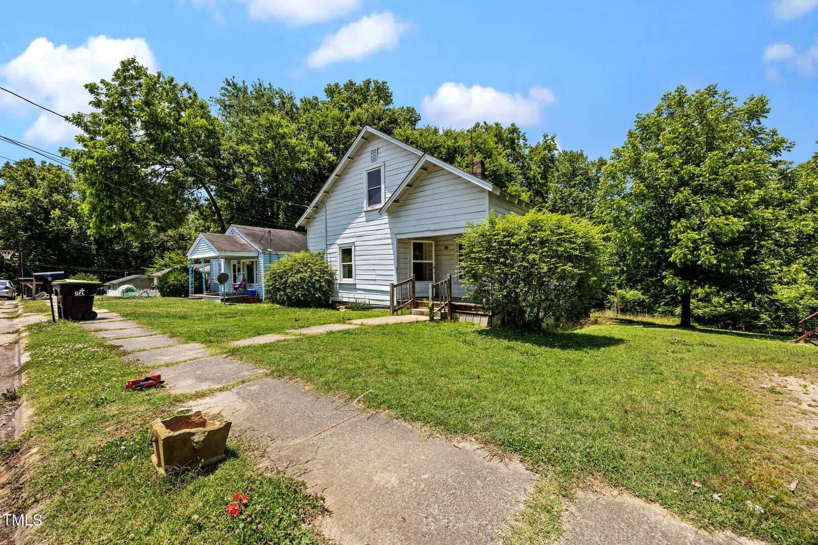 Two small white houses with front porches sit on a grassy lot with trees in the background. A cracked concrete driveway and scattered objects are visible in the foreground under a bright, blue sky.