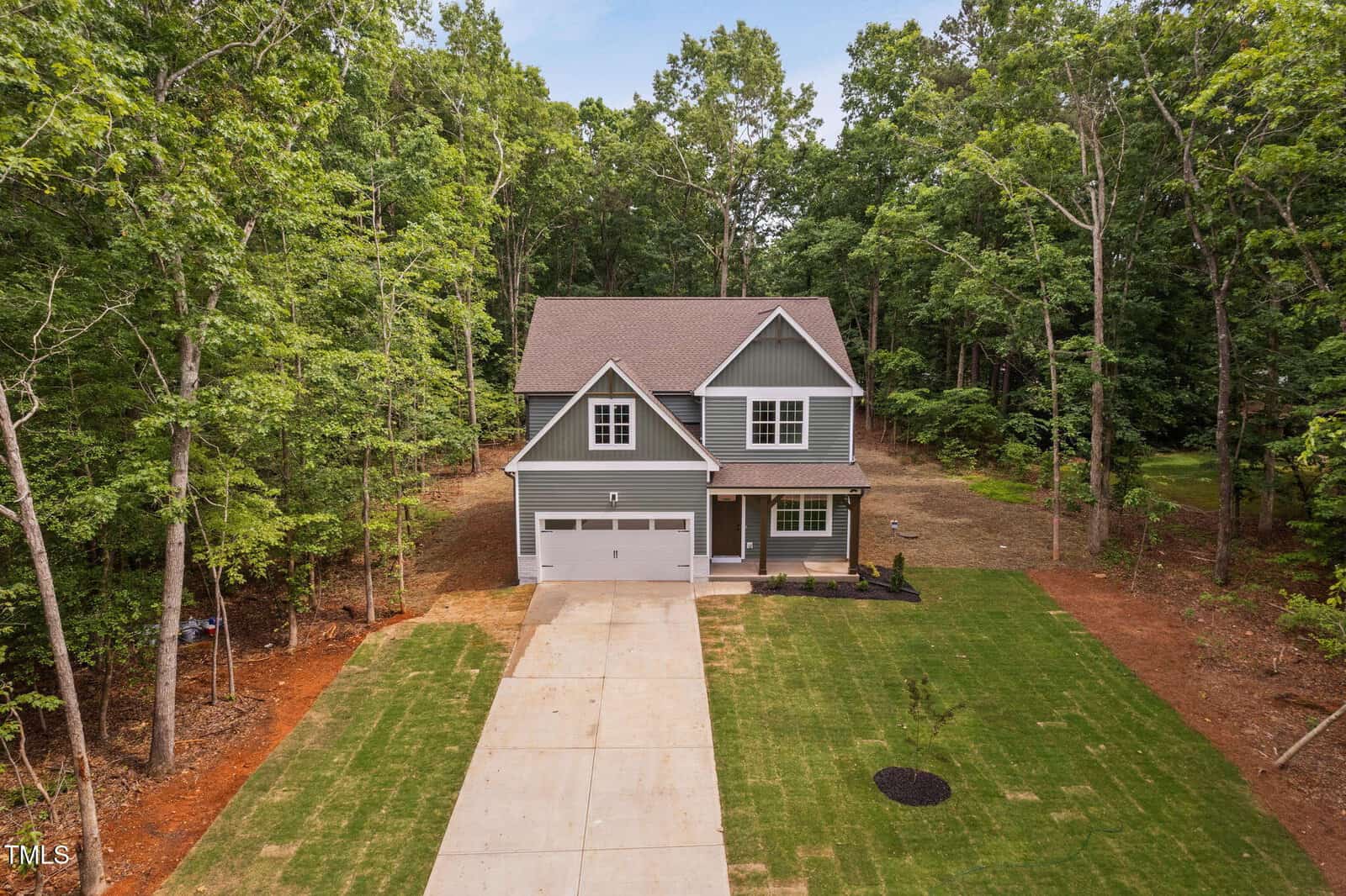 A two-story gray house with white trim and a double garage is surrounded by trees. The driveway is concrete, and the front yard has newly laid grass with a small circular patch of bare soil.