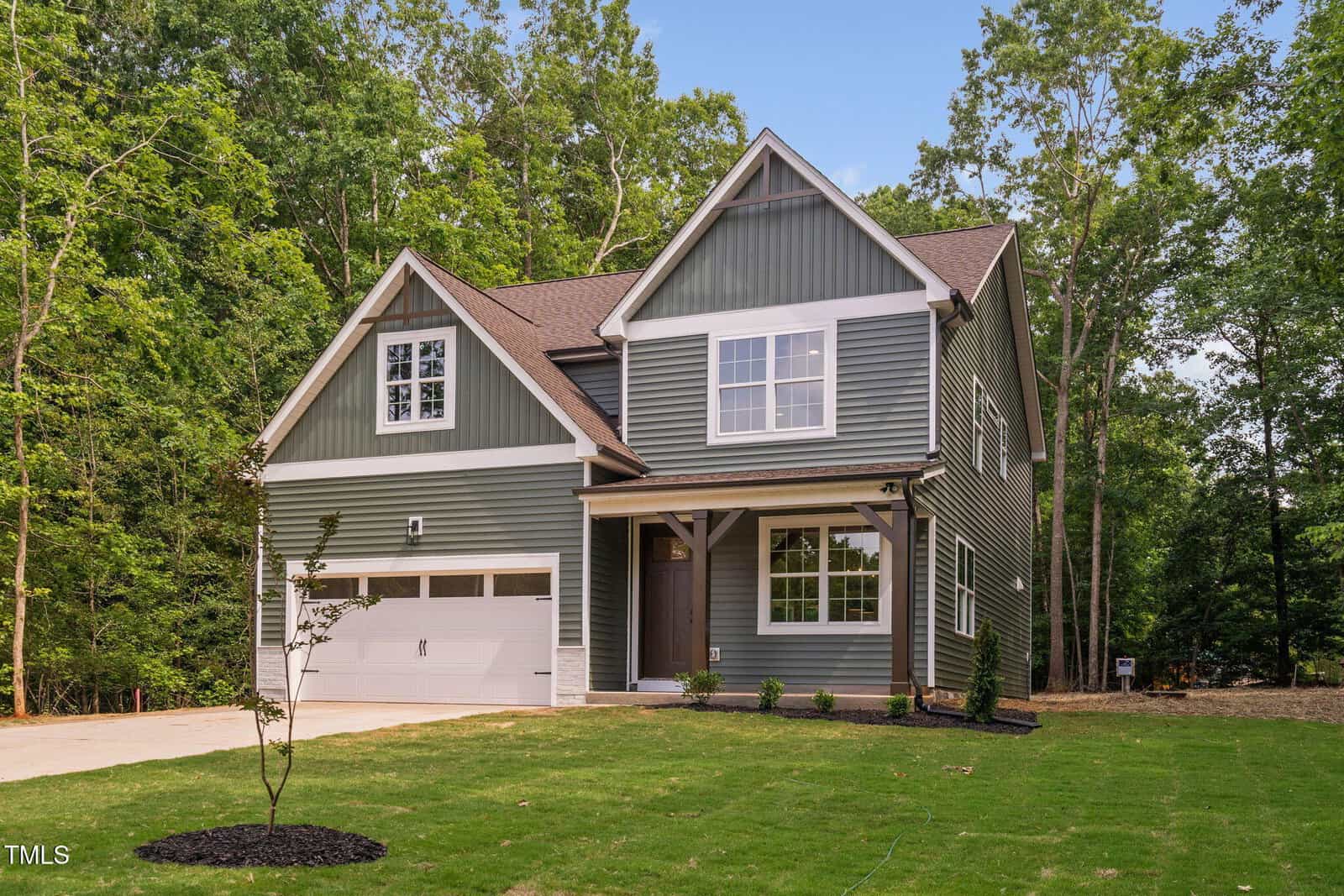Two-story gray house with white trim, gable roof, attached two-car garage, covered front porch, and a small tree planted in a green lawn. Surrounded by tall trees and bordered by a forested area.