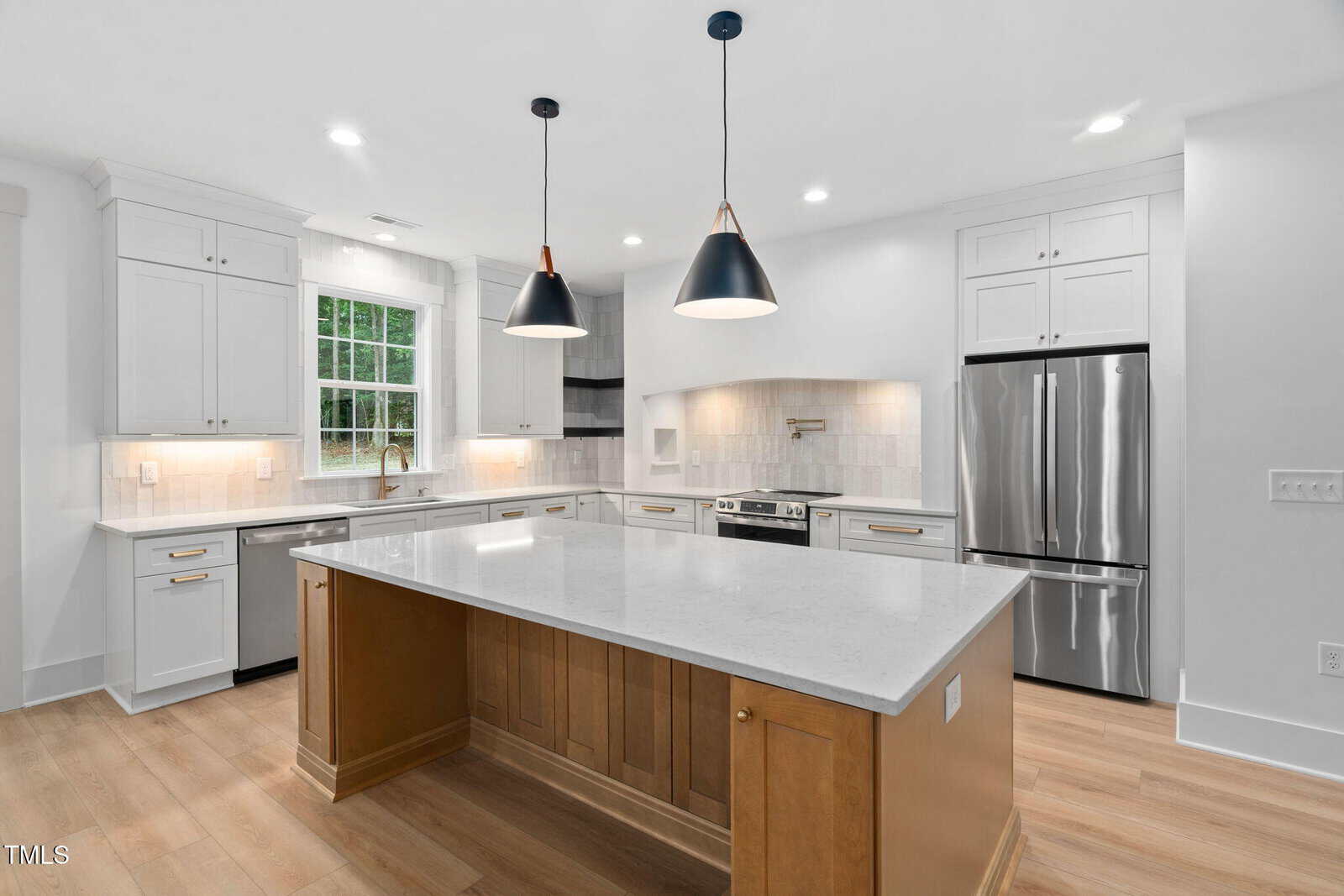 Modern kitchen at 114 Crow Drive, Louisburg with white cabinets, stainless steel appliances, a large island with a wood base, quartz countertops, pendant lighting, and light wood flooring. There is a window above the sink and recessed ceiling lights.