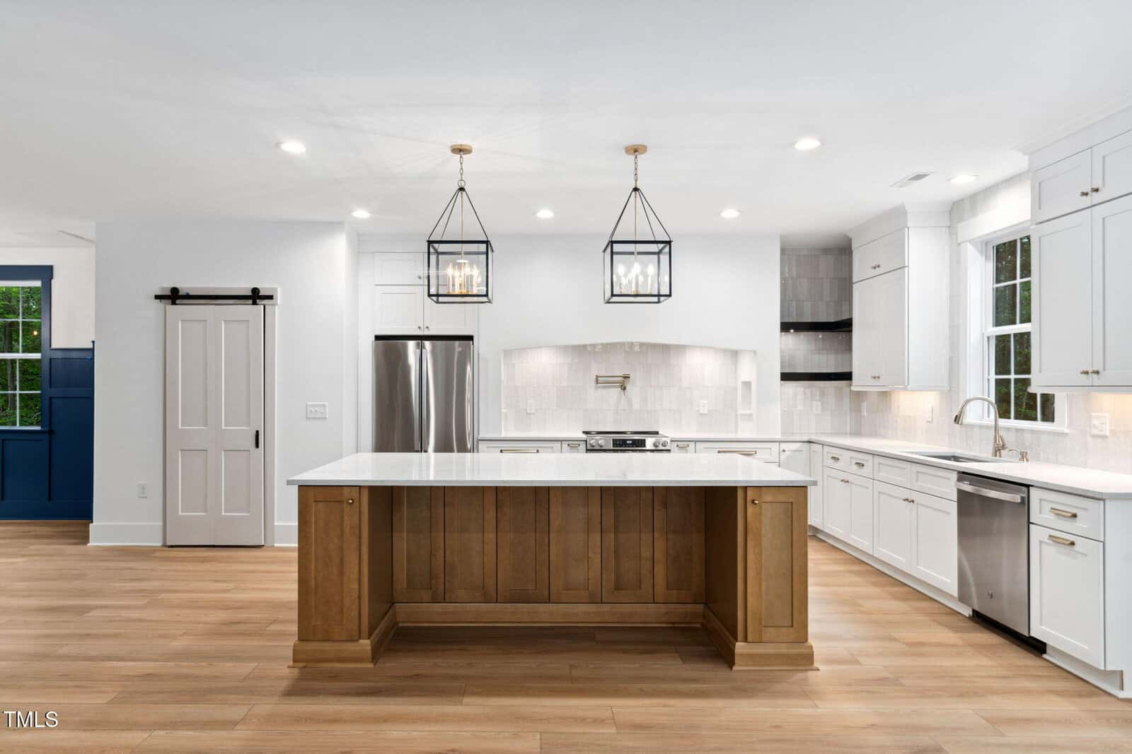 Modern kitchen at 125 Oswego Drive, Louisburg, featuring white cabinets, stainless steel appliances, a wood island with a white countertop, two pendant lights, light wood flooring, and a sliding barn door on the left. Large window above the sink.
