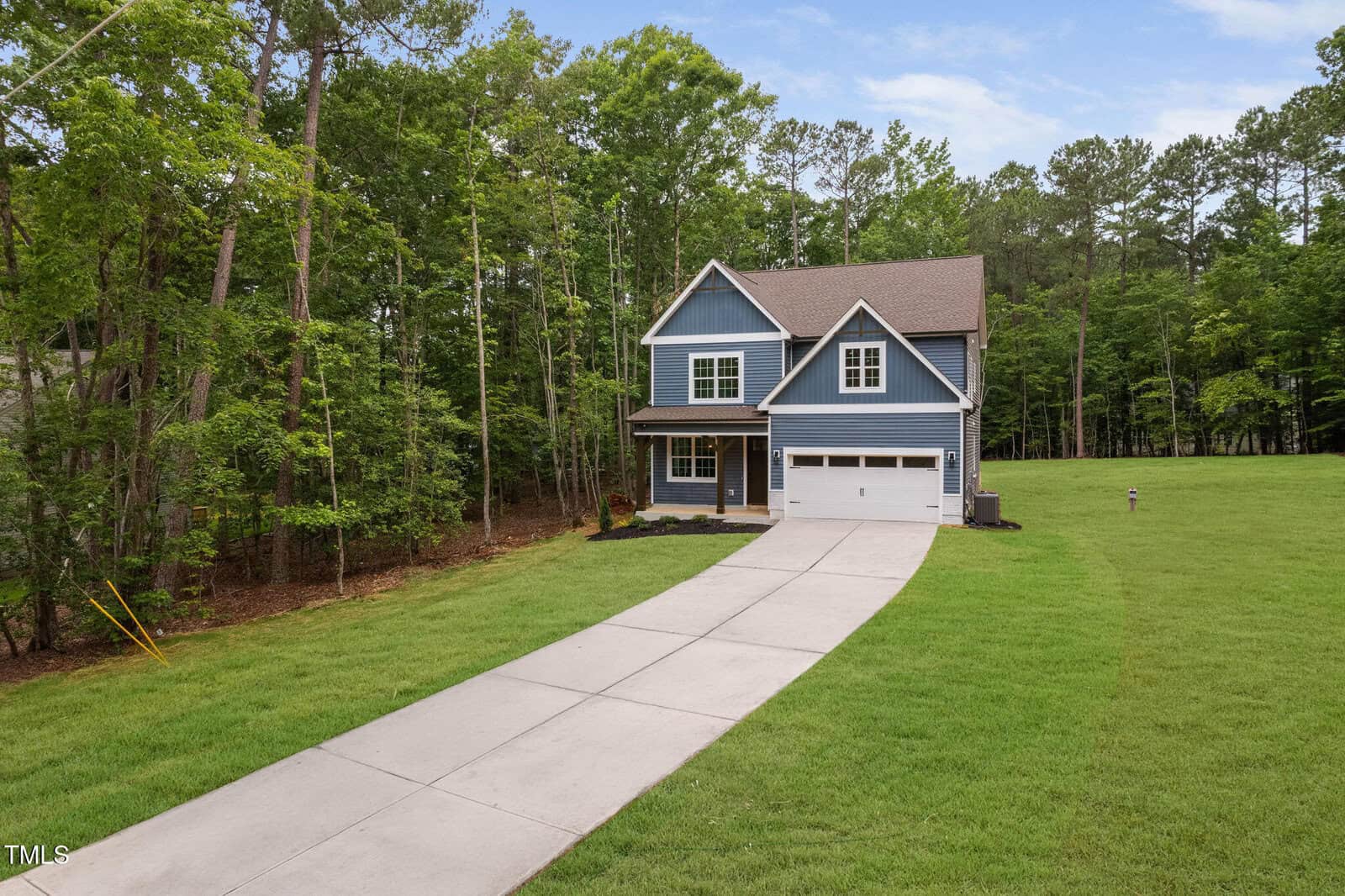 A two-story blue house with white trim and a double garage sits at the end of a long driveway, surrounded by green grass and trees under a partly cloudy sky.