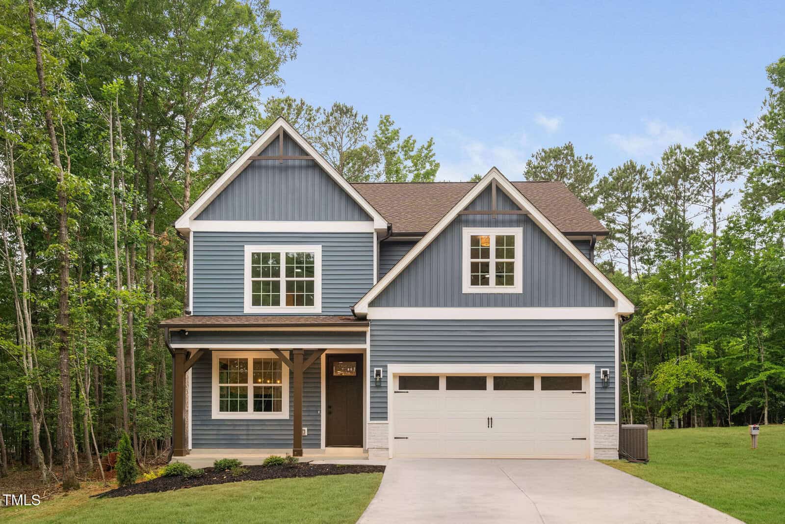 Two-story house with blue siding, white trim, and a covered front porch. Attached two-car garage and concrete driveway. Surrounded by tall trees and green grass. Sky is clear and blue.