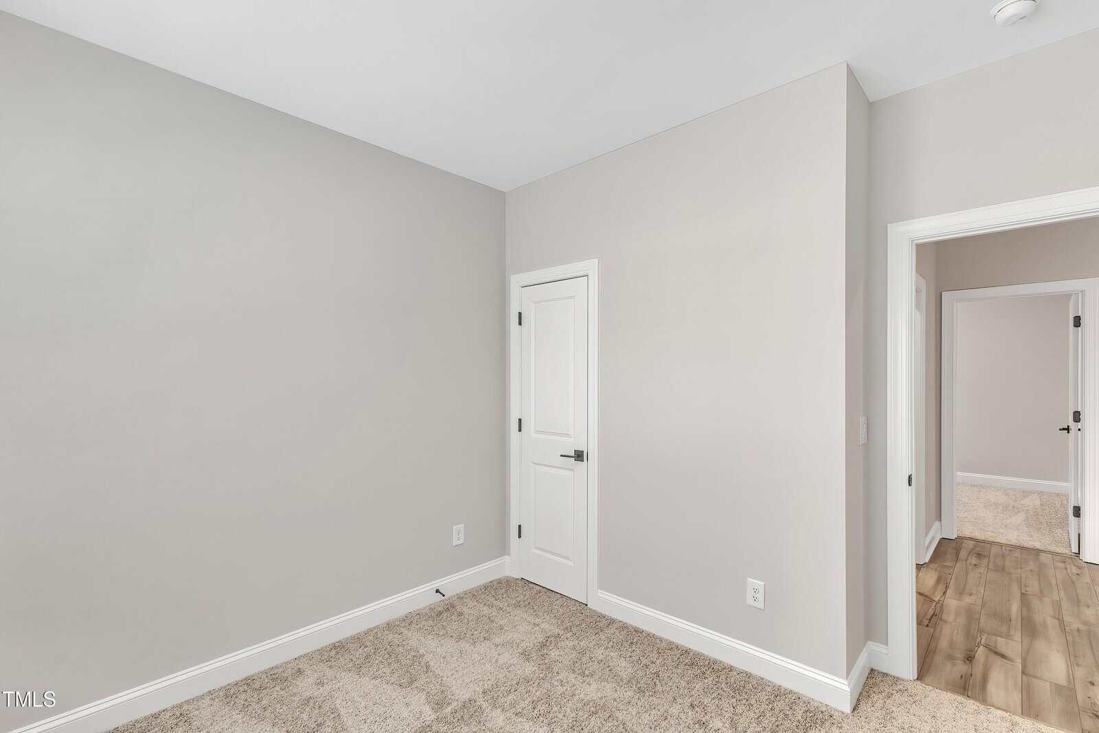A small, empty room with light beige walls, white trim, and a beige carpet. There is a closed white door in one corner, and an open doorway leading to a hall with wood flooring and another room visible.