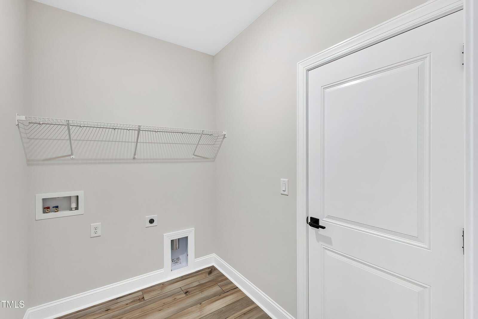 Laundry room with light gray walls, a wire shelf, utility hookups, an outlet, a small pet door near the floor, and a closed white door with a black handle. The floor features wood-style planks.