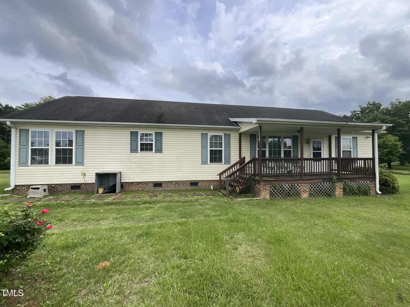 Charming single-story house at 2590 George Winston Road, Bullock, featuring light yellow siding, a black roof, blue shutters, and a covered front porch with wooden steps. The grassy lawn is adorned with bushes and flowers in the foreground.