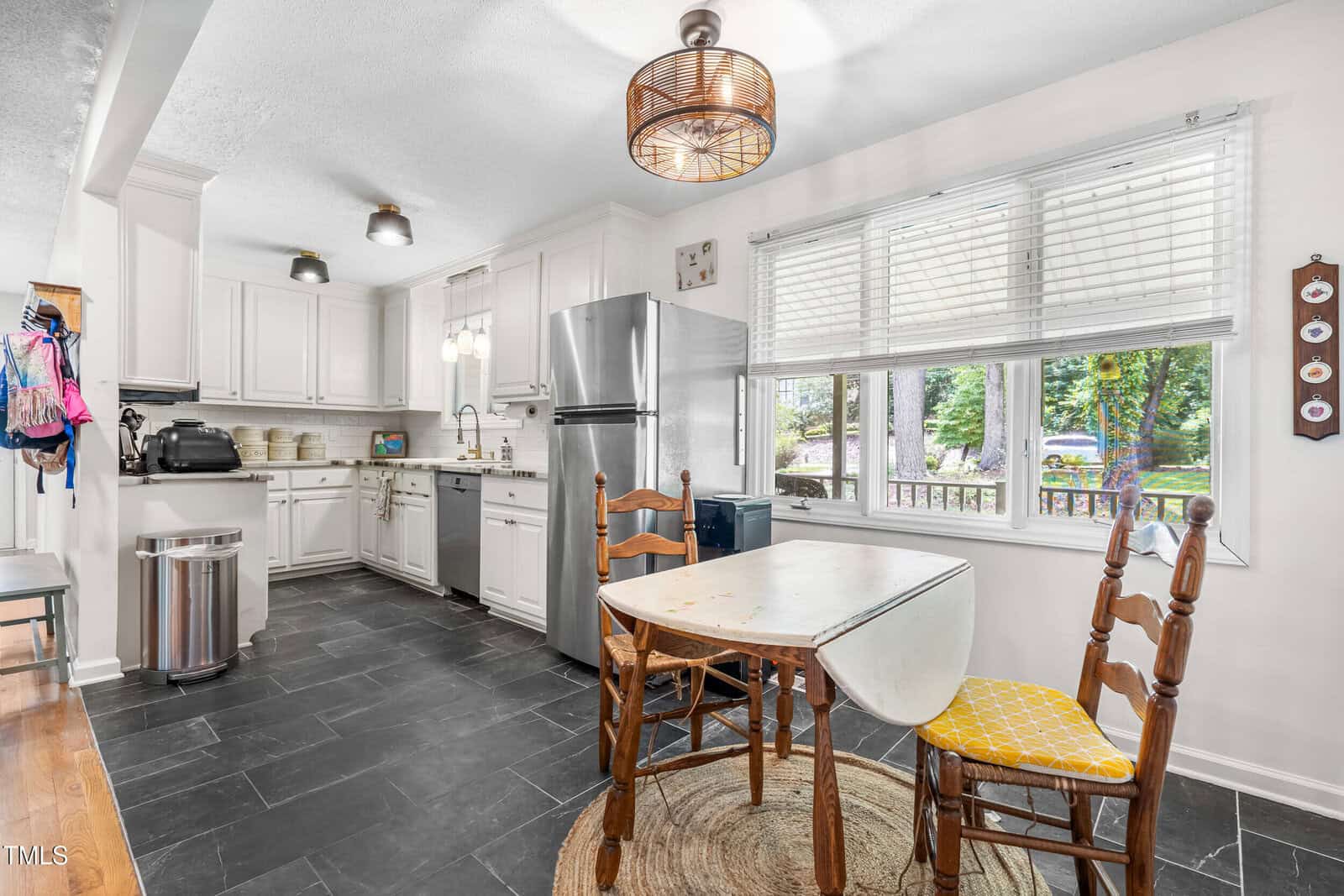 A bright kitchen with white cabinets, stainless steel appliances, a small dining table with two wooden chairs, black tile floor, and large window letting in natural light.