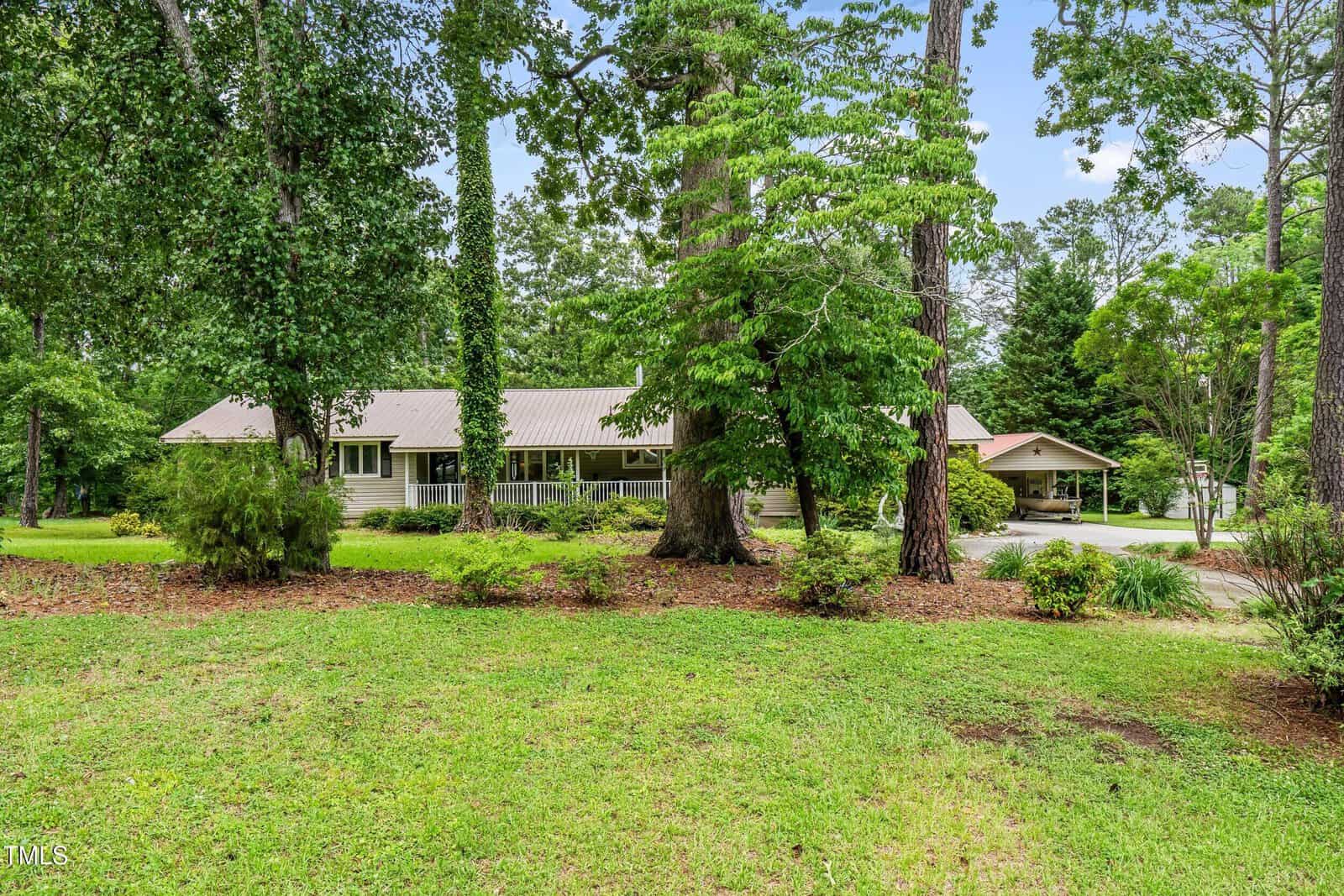 A single-story house with a metal roof sits behind several tall trees and shrubs, surrounded by green grass and landscaping. A driveway leads to a carport on the right side of the house.