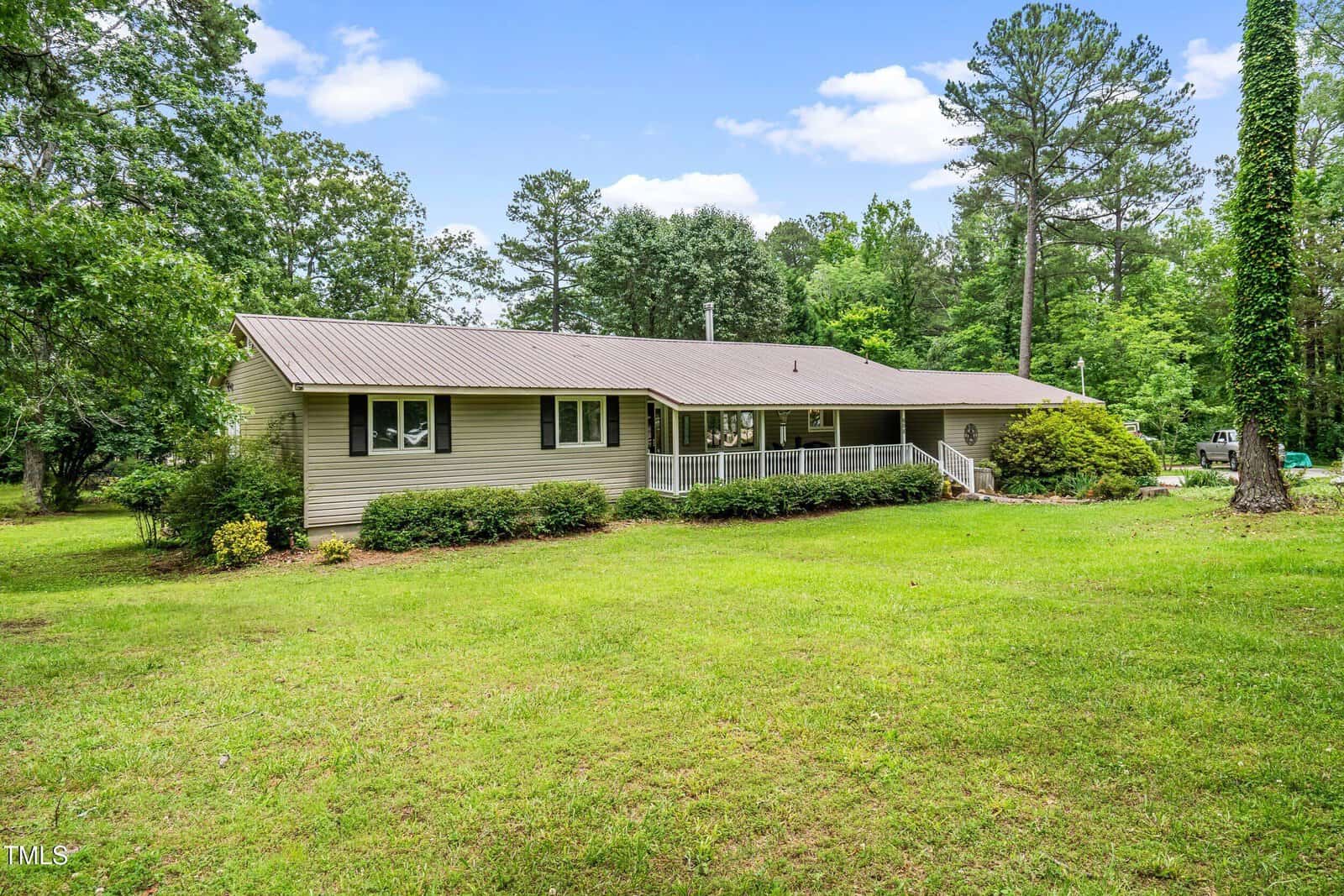 Charming single-story beige house at 608 20th Street, Butner, with a metal roof and front porch, nestled among green grass, shrubs, and tall trees under a partly cloudy sky.