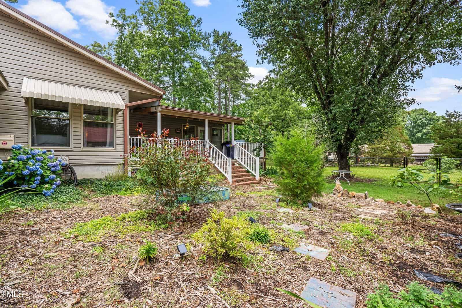 A single-story house with a covered porch, brick steps, and metal railings overlooks a mulched garden with various small plants and shrubs. Large trees provide shade, and a green lawn extends into the background.