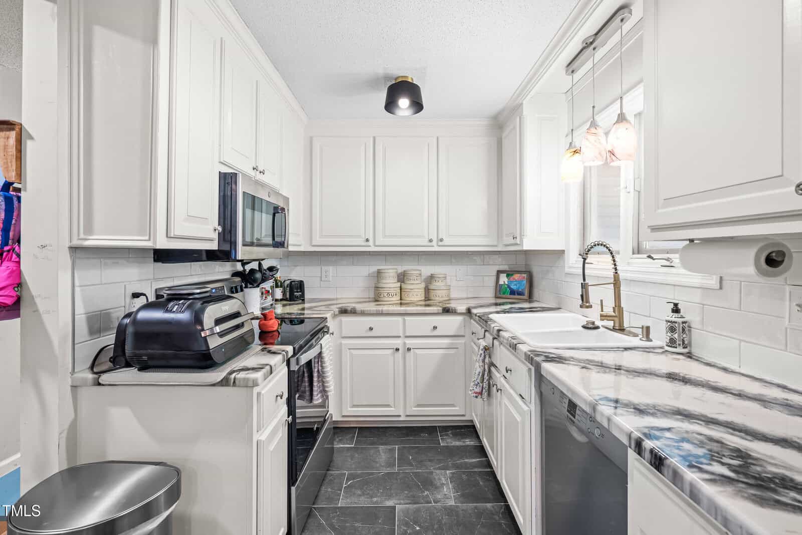 A modern kitchen with white cabinets, stainless steel appliances, a marble-patterned countertop, a double sink, and black tiled floors. Pendant lights hang over the sink, and small appliances are on the counters.