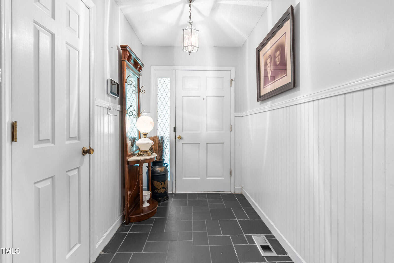 A small entryway with white walls and wainscoting, black tile flooring, a white front door, a side table with a lamp and decor, a framed photo on the wall, and a ceiling light fixture.