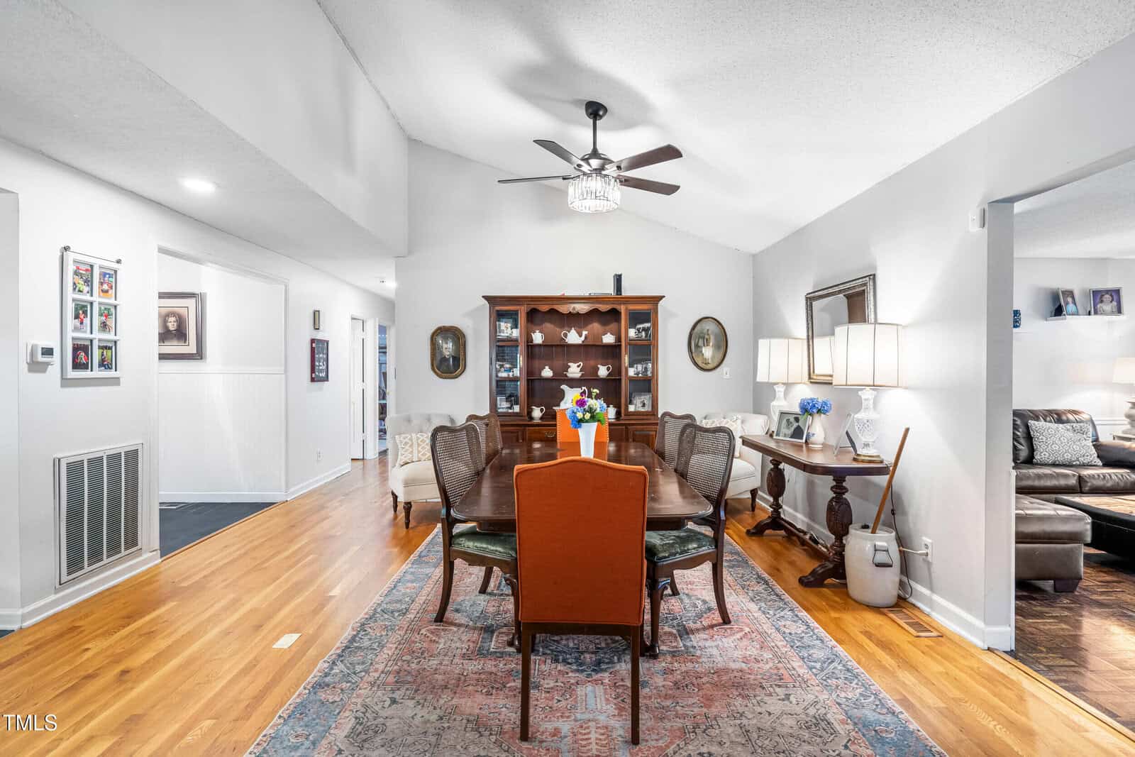 Dining room with hardwood floors, large rug, wooden table and chairs, china cabinet, sideboard, and ceiling fan; hallway and living room visible in background. Flowers and lamps decorate the space.