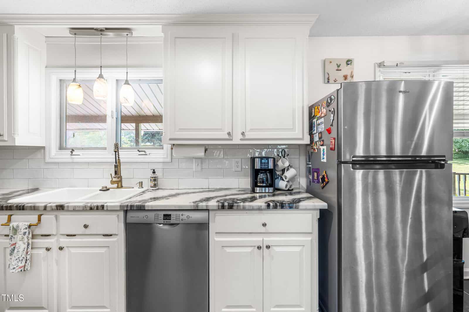 A kitchen with white cabinets, a marble countertop, stainless steel dishwasher, and refrigerator. A sink is under a window, and various magnets and items are on the fridge. Pendant lights hang above the sink.