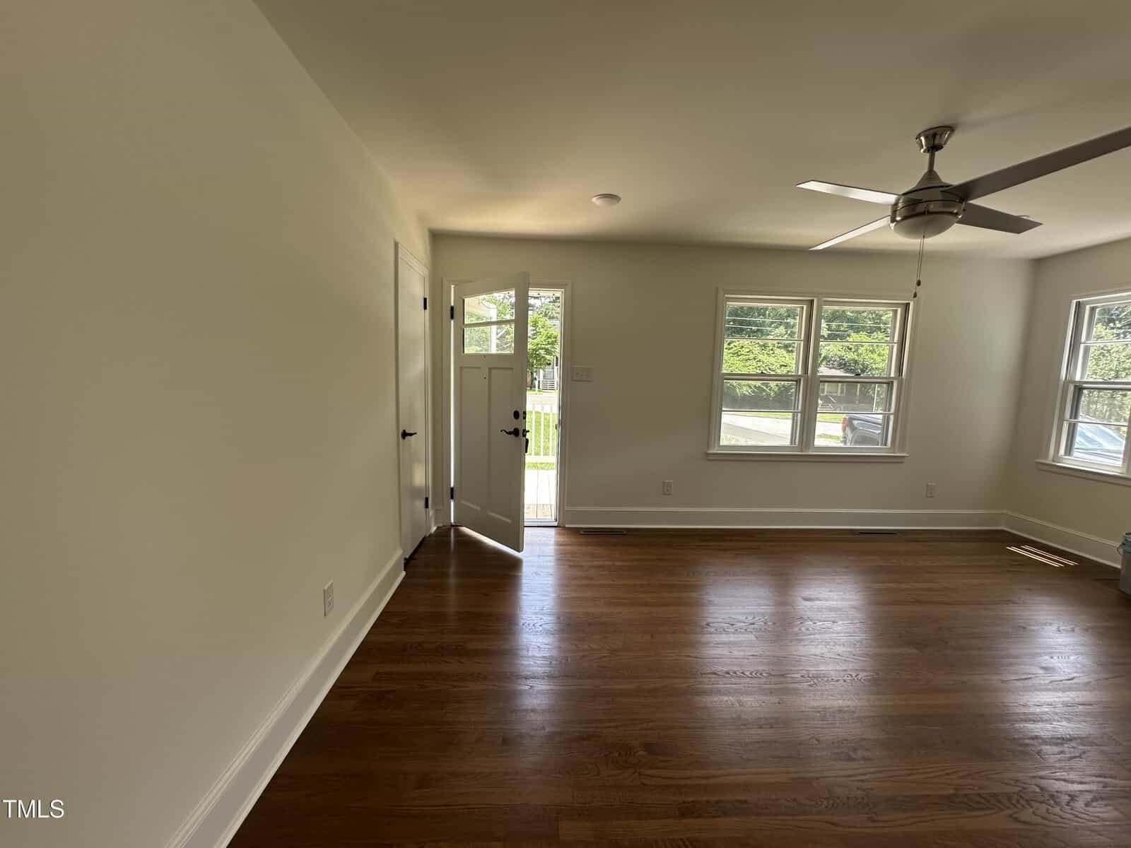 A room with hardwood floors, white walls, a ceiling fan, three large windows, and a door that is slightly open to the outside. Sunlight streams in, illuminating the space.