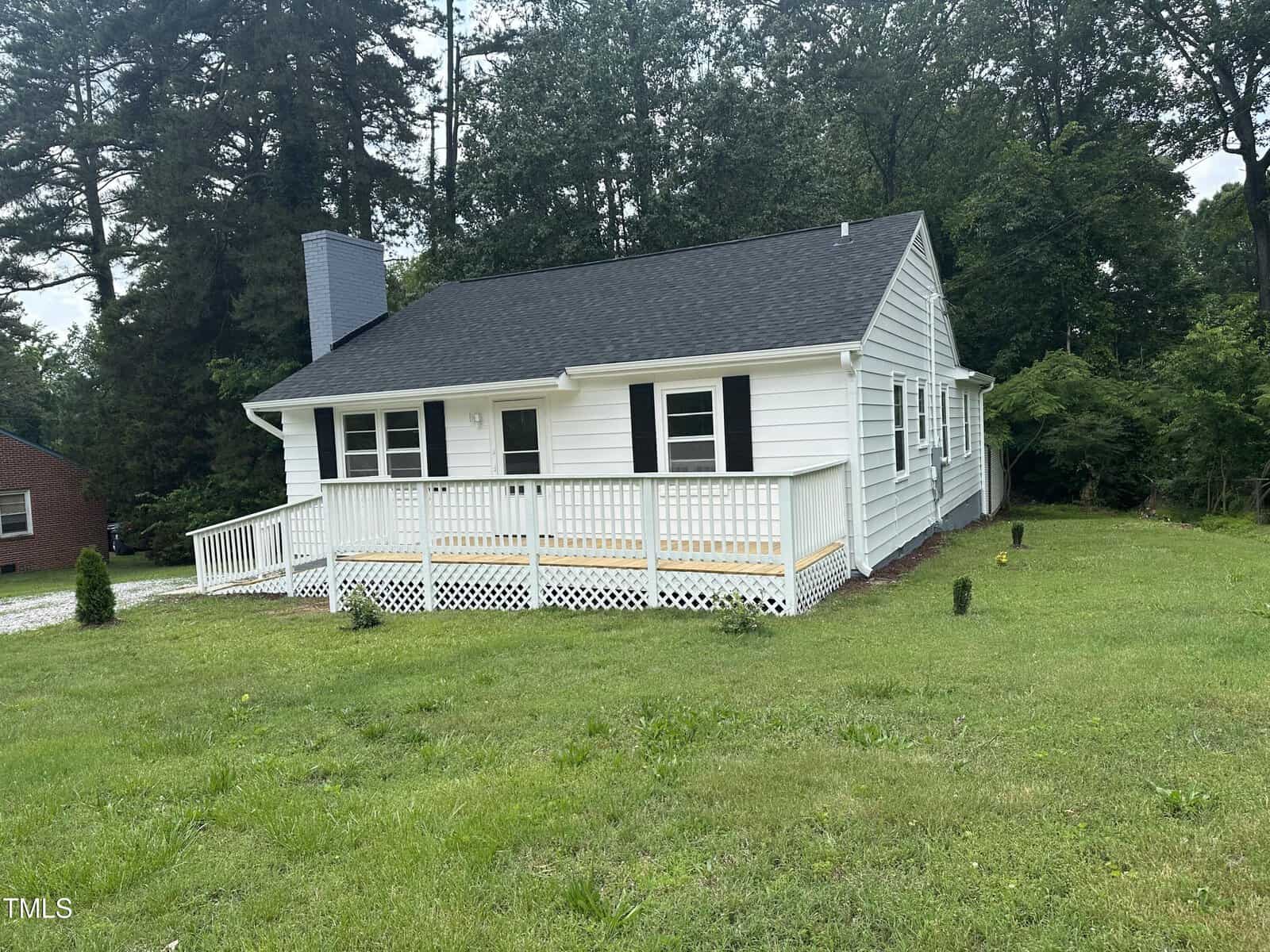 A small, single-story white house with black shutters and a dark shingled roof sits on a grassy lawn bordered by trees at 902 Roanoke Avenue in Henderson, featuring a front deck with classic white railings.