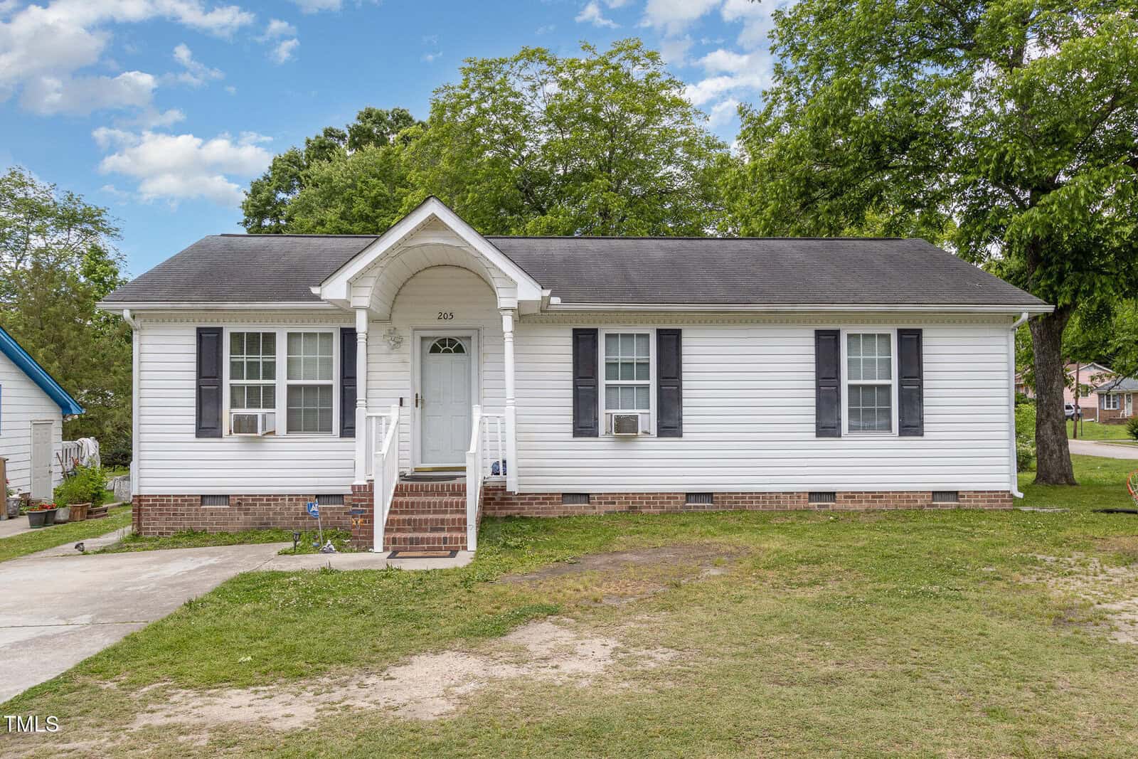 Charming single-story white house with black shutters at 205 Person Street, Oxford. Features a small front porch with steps, two window air conditioning units, patchy grass yard, paved driveway on the left, trees, and nearby houses.