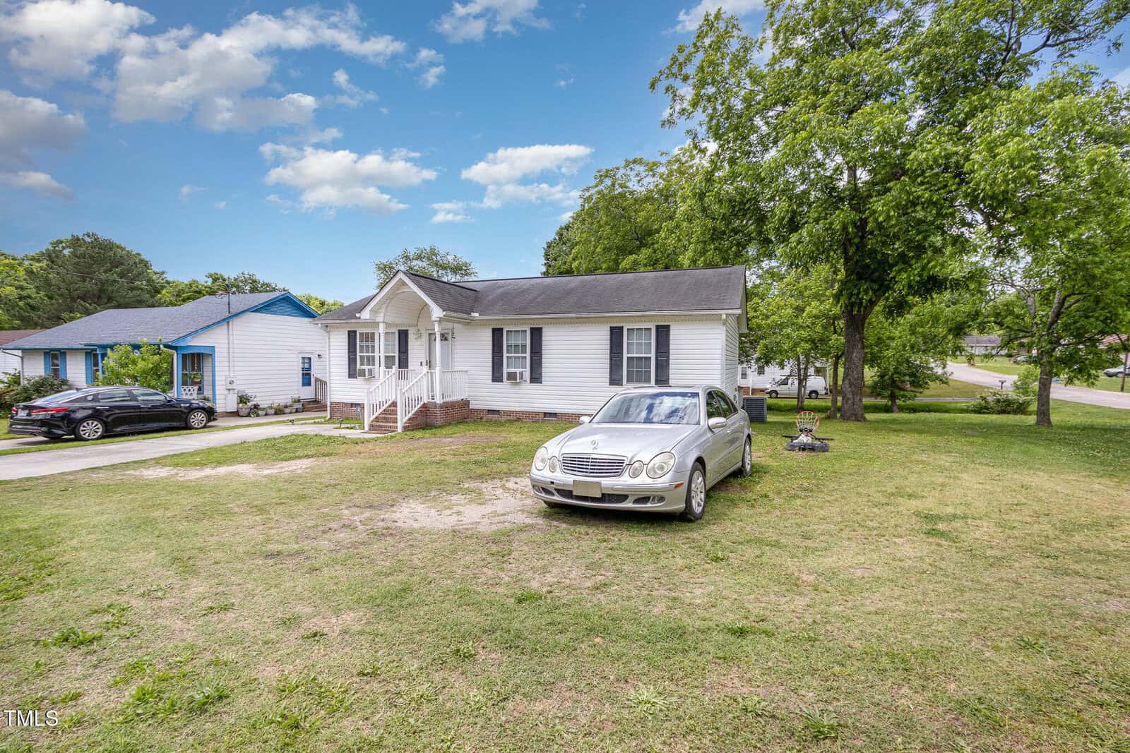 Single-story white house with a front porch, black shutters, and a gray roof. A silver car is parked on the lawn, and trees surround the property with another house visible next door under a partly cloudy sky.