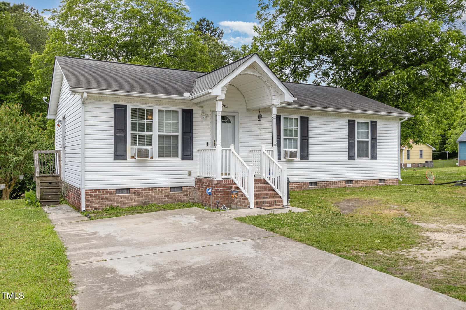 A single-story white house with black shutters and a gray shingle roof, featuring a small front porch with white railings, brick steps, two window air conditioners, and a concrete driveway. Trees and grass surround the house.