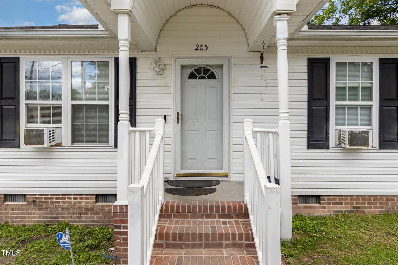 A white house with a covered front porch, brick steps, white railings, black shutters, and two window air conditioning units. The front door is centered under an arched portico, with the number 205 above it.